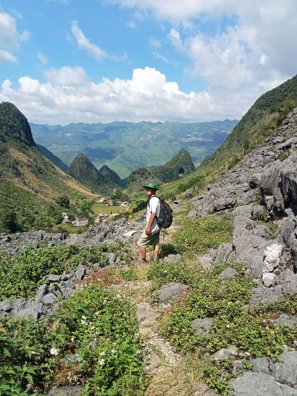 A walker with a light daypack on a rocky limestone path above a cultivated valley on the Dong Van Karst Plateau.