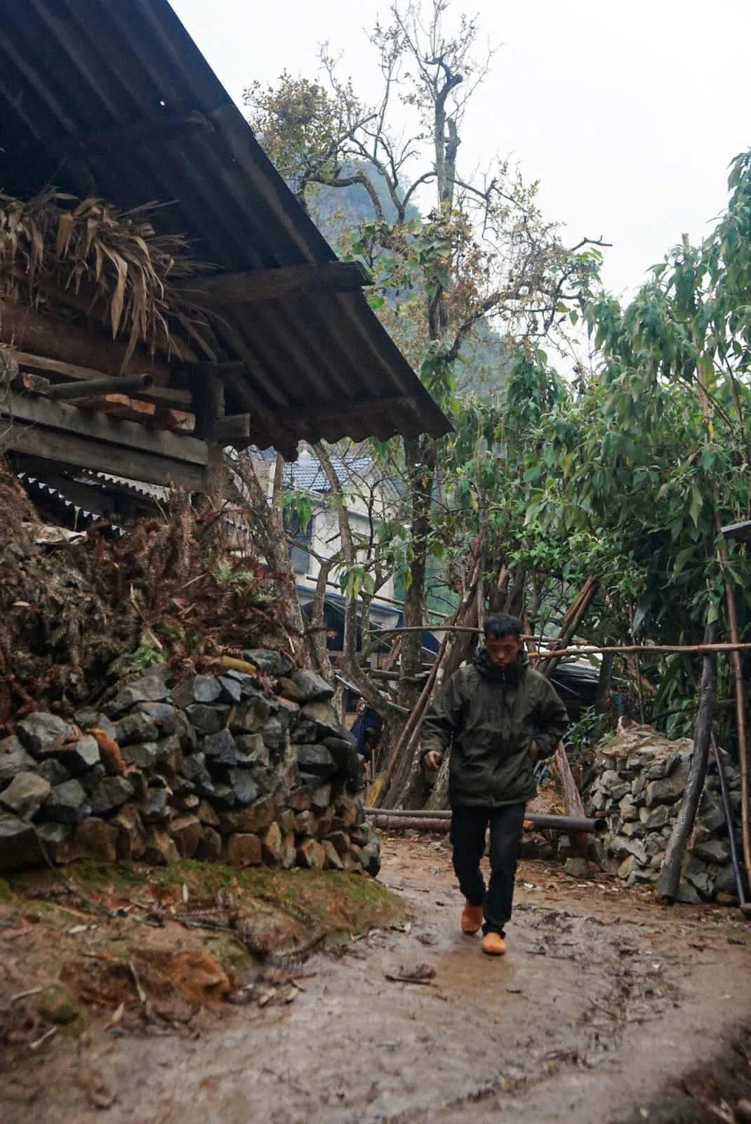 A walker on a wet, muddy village path between dry-stone walls and wooden houses in the Dong Van region.