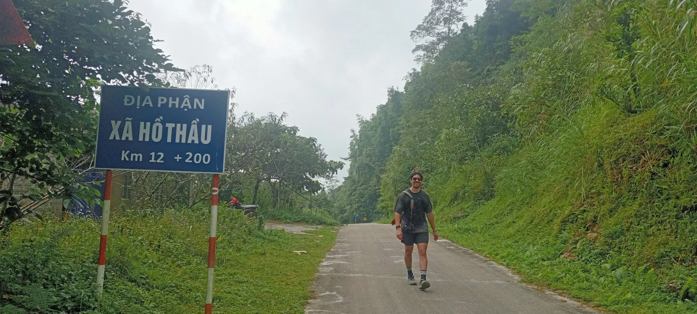 A trekker walks past a road sign for Hồ Thầu commune on a paved mountain road during the Ho Thau to Nam Ai stage, Hoang Su Phi.