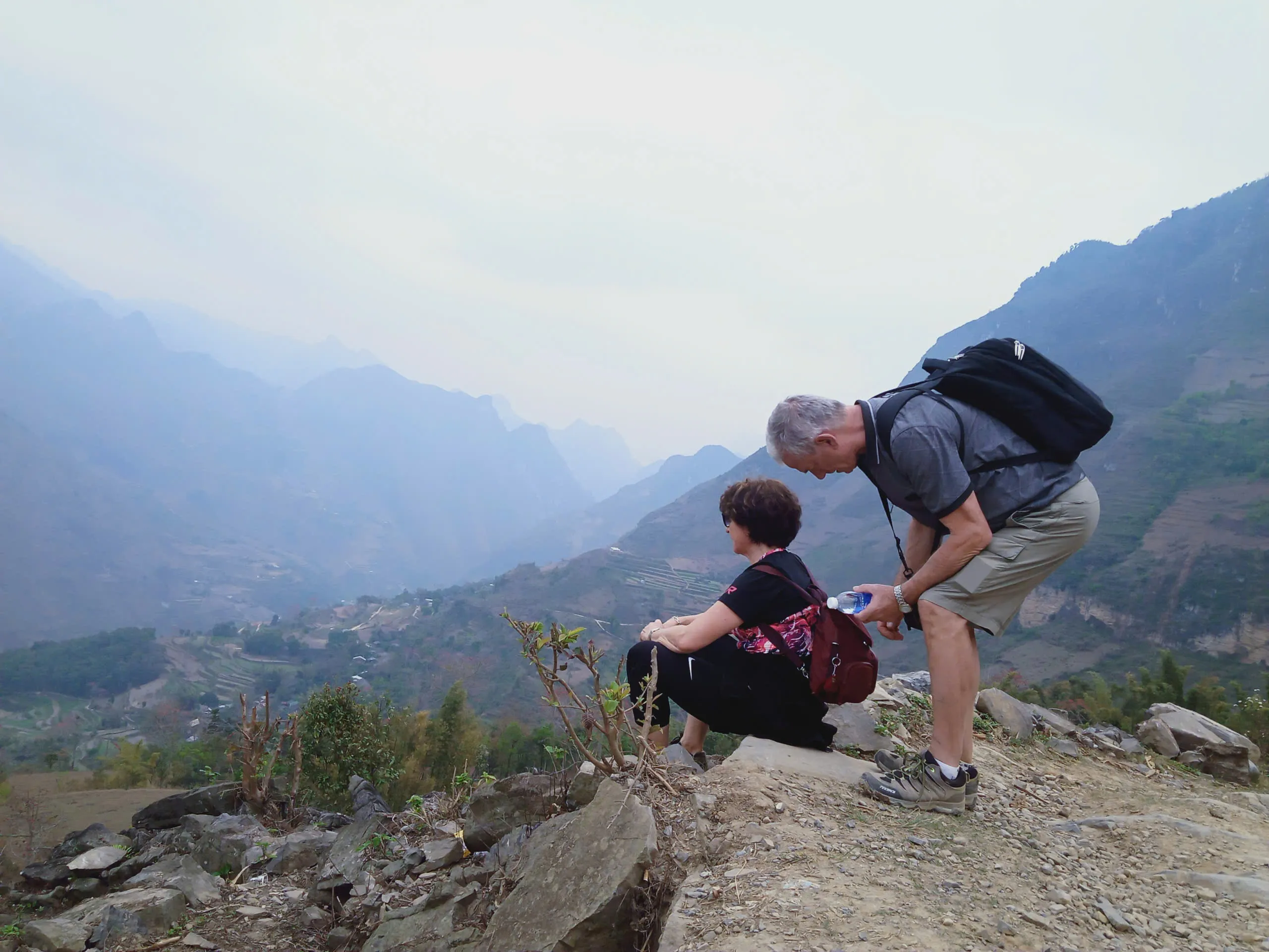 Hikers with daypacks resting on a rocky ledge above a misty limestone canyon in eastern Ha Giang.
