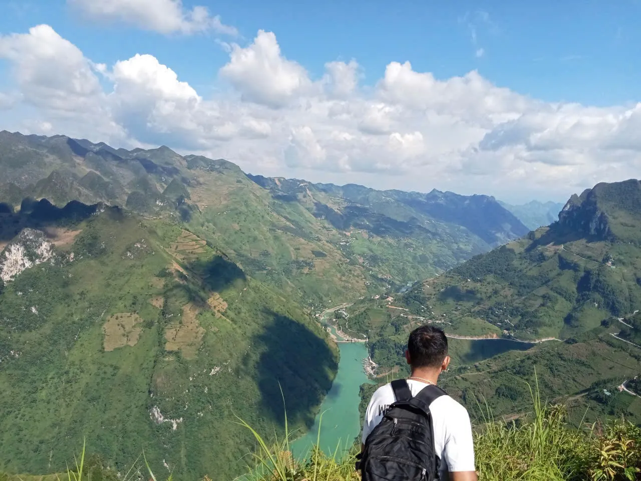 A walker with a daypack looking over a turquoise river and farmed slopes from a mountain rim in the Ma Pi Leng area.