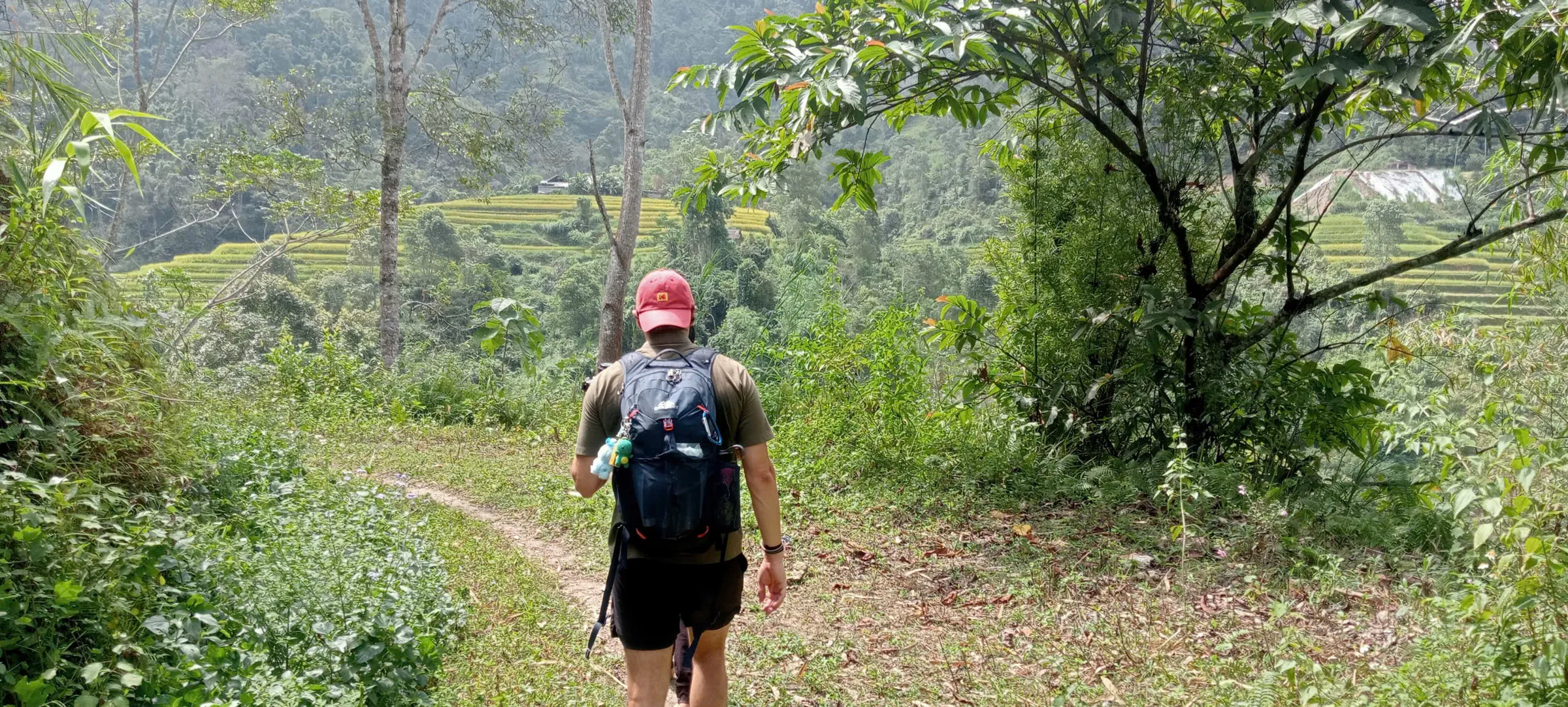 A trekker with a daypack walks a narrow dirt path above terraced slopes on the Nam Ai to Phin Ho stage in Hoang Su Phi