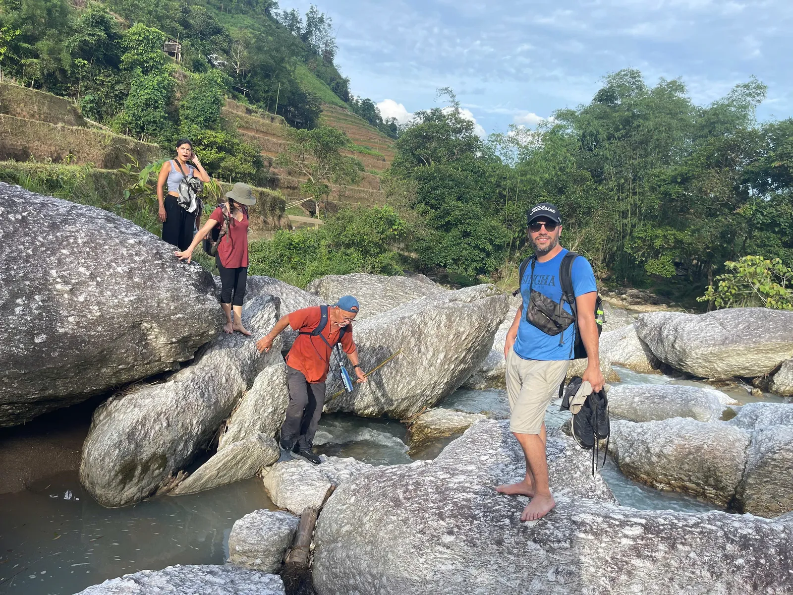 A guide and trekkers cross boulders beside a shallow stream below terraced slopes near Nam Hong, Hoang Su Phi.